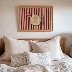 Puede incluir: Una obra de arte enmarcada que representa un plato de tostadas con mantequilla, un cuchillo y una tapa de mermelada a cuadros rojos y blancos sobre un fondo a rayas rosas y marrones. La obra de arte est&aacute; sobre una cama con almohadas blancas y cojines estampados florales.