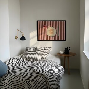 Puede incluir: Una habitaci&oacute;n con una cama con ropa de cama a rayas blancas y negras y una almohada a juego. Una obra de arte enmarcada que representa el desayuno cuelga sobre la cama. Una peque&ntilde;a mesa auxiliar de madera con una jarra negra y una taza blanca.