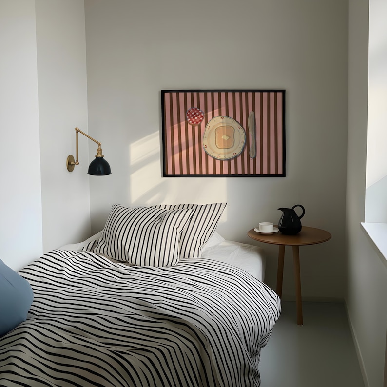 Puede incluir: Una habitaci&oacute;n con una cama con ropa de cama a rayas blancas y negras y una almohada a juego. Una obra de arte enmarcada que representa el desayuno cuelga sobre la cama. Una peque&ntilde;a mesa auxiliar de madera con una jarra negra y una taza blanca.
