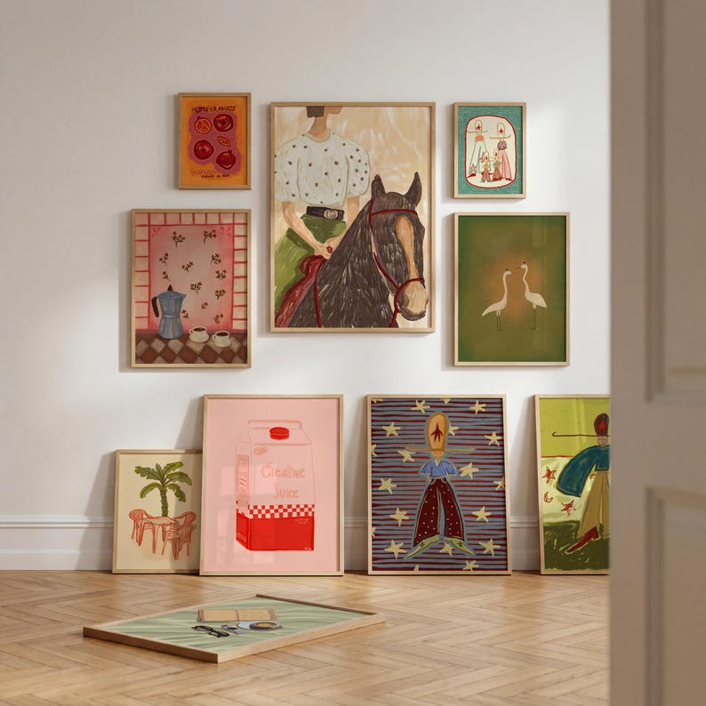 May include: A gallery wall arrangement featuring nine framed art prints. The collection includes illustrations of a person on a horse, a coffee pot, two cranes, a carton labeled Creative Juice, a star-patterned figure, and a palm tree scene.