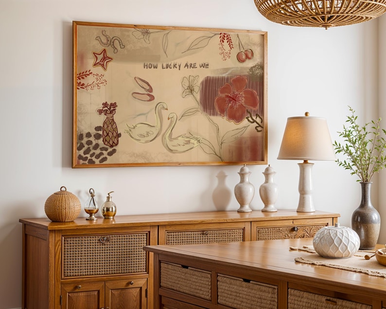 Puede incluir: Una obra de arte enmarcada con el texto "HOW LUCKY ARE WE" presenta cisnes esbozados, flores y otras ilustraciones en tonos rojos y marrones. La obra de arte cuelga sobre un mueble de madera con objetos decorativos.