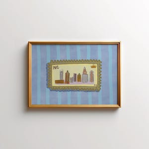 May include: A framed artwork depicting a cityscape in a postage stamp design. The stamp, with the letters "NC" and a crown, is set against a blue striped background. The frame is gold-coloured.