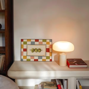 Op de afbeelding: Een ingelijst kunstwerk met drie groene olijven aan een spies, tegen een crèmekleurige achtergrond. De lijst heeft een kleurrijk dambordpatroon. Een witte paddenstoelvormige lamp en een rood boek staan op een plank.