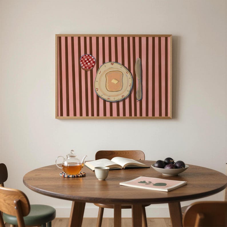 Puede incluir: Una pintura enmarcada representa una escena de desayuno con una rebanada de tostada con mantequilla, un tarro de mermelada y un cuchillo sobre un fondo a rayas rosas y marrones. Una mesa redonda de madera con tetera, libro, taza y un cuenco de ciruelas.
