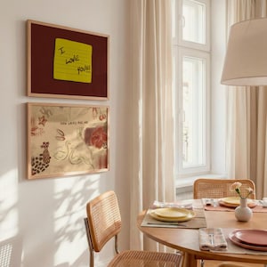 Puede incluir: Dos impresiones art&iacute;sticas enmarcadas cuelgan de una pared blanca. La impresi&oacute;n superior tiene un fondo granate con una nota adhesiva amarilla que dice "I LOVE YOU!!!". La impresi&oacute;n inferior tiene un fondo dorado con un dise&ntilde;o floral y de cisnes y el texto "HOW LUCKY ARE WE".