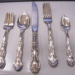 May include: A set of five silver-colored flatware pieces: two forks, two spoons, and a knife. Each piece has a decorative, ornate handle. The knife is in the center, flanked by a spoon and a fork on each side. The silverware is arranged on a light-colored surface.