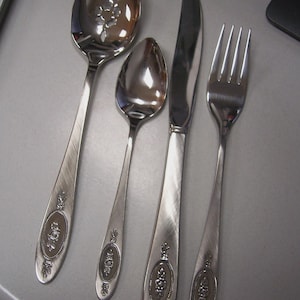 May include: A set of silver-colored flatware including a slotted spoon, a spoon, a knife, and a fork. Each piece has a decorative floral design on the handle.