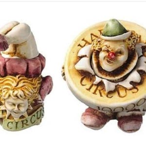 May include: Two whimsical clown figurines. One is upside down with a white hand and a red and pink ruffled collar. The other is a clown face emerging from a yellow coin that reads "HAPPY CIRCUS".