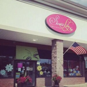 Puede incluir: Vista exterior de una tienda llamada "Sierra Lily". La tienda tiene un letrero ovalado rosa con letras blancas. Una bandera estadounidense ondea al viento. La entrada de la tienda est&aacute; decorada con flores y dise&ntilde;os florales.