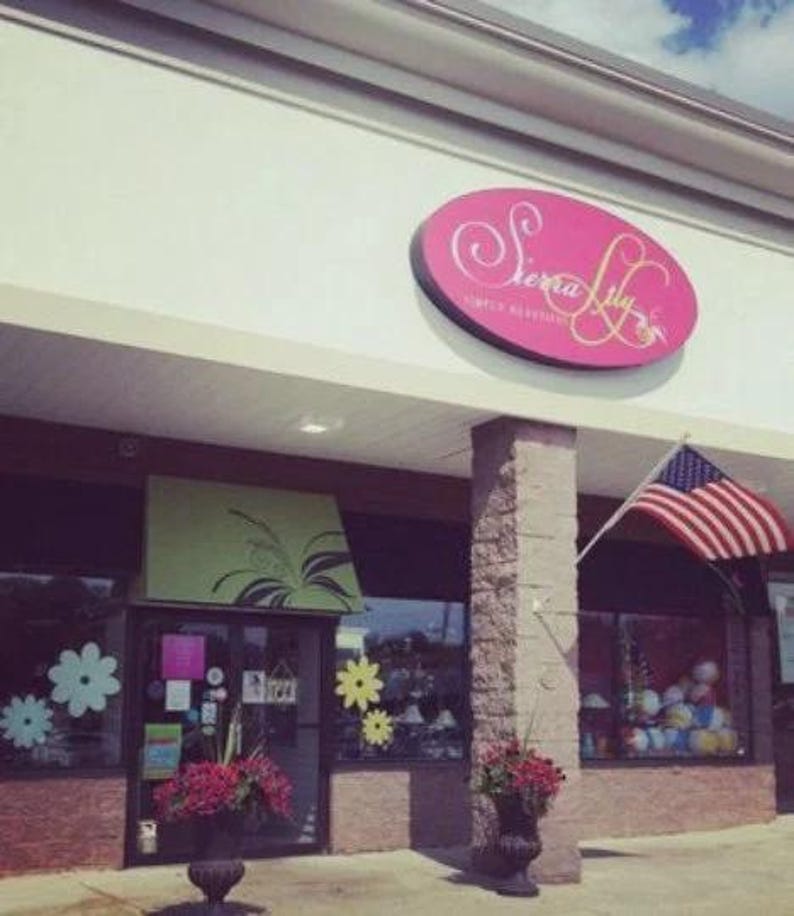 Puede incluir: Vista exterior de una tienda llamada "Sierra Lily". La tienda tiene un letrero ovalado rosa con letras blancas. Una bandera estadounidense ondea al viento. La entrada de la tienda est&aacute; decorada con flores y dise&ntilde;os florales.