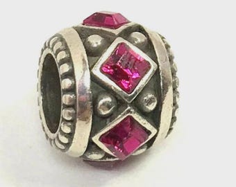 May include: Silver-toned bead with a central design featuring three fuchsia-colored square gemstones. The bead has a cylindrical shape with a textured surface and is likely used for jewelry making.