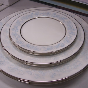 May include: A stack of white porcelain plates with a light blue floral pattern and silver trim. The set includes plates of varying sizes, creating a layered effect. The plates are clean and appear unused, suitable for formal dining.