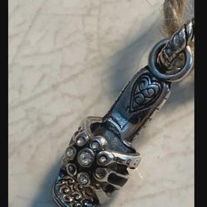 Puede incluir: Un dije plateado con forma de zapato de tac&oacute;n alto, adornado con piedras preciosas transparentes y un dise&ntilde;o de coraz&oacute;n decorativo. El dije est&aacute; sujeto a una cuerda trenzada, lo que sugiere que podr&iacute;a ser un colgante o un elemento decorativo para joyer&iacute;a.