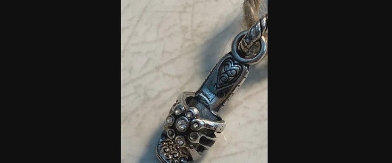 Puede incluir: Un dije plateado con forma de zapato de tac&oacute;n alto, adornado con piedras preciosas transparentes y un dise&ntilde;o de coraz&oacute;n decorativo. El dije est&aacute; sujeto a una cuerda trenzada, lo que sugiere que podr&iacute;a ser un colgante o un elemento decorativo para joyer&iacute;a.