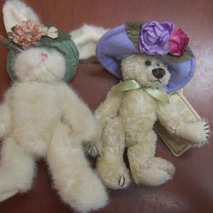 May include: Two plush toys: a bunny and a teddy bear. The bunny wears a green hat with pink flowers, and the bear wears a purple hat with pink and purple flowers. Both toys are cream-colored and have black eyes and noses.