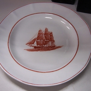 May include: White ceramic plate with a red border and a detailed illustration of a sailing ship in the center. The ship is depicted with multiple sails and is sailing on the water. A decorative plate for nautical decor.