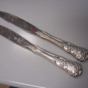 May include: Two silver butter knives with ornate floral detailing on the handles. The blades are smooth and reflective, and the handles have a raised floral pattern. The knives are likely part of a vintage silverware set.