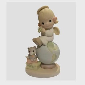 May include: A ceramic figurine featuring a seated angel with wings, holding a small object, atop a globe. A small cat figurine sits on a stack of boxes. The color palette includes soft pastels of beige, white, and light green.