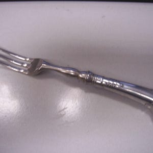 May include: A silver fork with three tines and a decorative handle. The handle has a curved, scroll-like design at the end and a detailed pattern near the fork's head. The fork is made of a shiny, reflective metal.