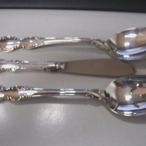 May include: Three pieces of silverware: two spoons and a butter knife. The silverware is silver-colored with ornate, floral-like detailing on the handles. The spoons have a rounded bowl shape, and the knife has a flat, rounded blade.