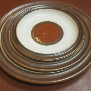 May include: A stack of ceramic plates with a brown and white color scheme. The plates have a concentric design, with a brown outer rim, a white inner section, and a reddish-brown center. The plates are stacked on top of each other.