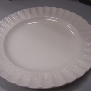 May include: Off-white, oval-shaped serving platter. The rim features a scalloped edge with a textured, embossed pattern. The center of the platter is smooth and glossy. A classic piece for serving food.