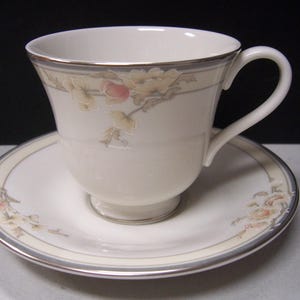 May include: Cream-colored teacup and saucer set with floral and gold trim. The cup features a curved handle and a delicate floral pattern with pink and yellow accents. The saucer mirrors the cup's design, creating a cohesive and elegant look.