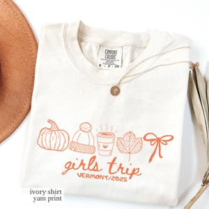 May include: Ivory-colored t-shirt with a pumpkin, hat, coffee cup, leaf, and bow graphic in orange. The text "girls trip VERMONT/2025" is printed below the graphics. A gold necklace is draped across the shirt.