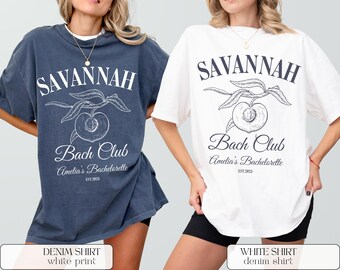 Può includere: Due magliette oversize, una blu denim e una bianca, ciascuna con un design grafico. Il design include la parola "SAVANNAH", un'illustrazione di pesca e il testo "Bach Club, Amelia's Bachelorette, EST. 2025".