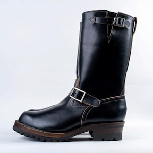May include: Black leather motorcycle boot with a tall shaft, featuring two adjustable buckle straps. The boot has a lug sole and a stacked heel, with visible stitching details. The overall design is rugged and durable.