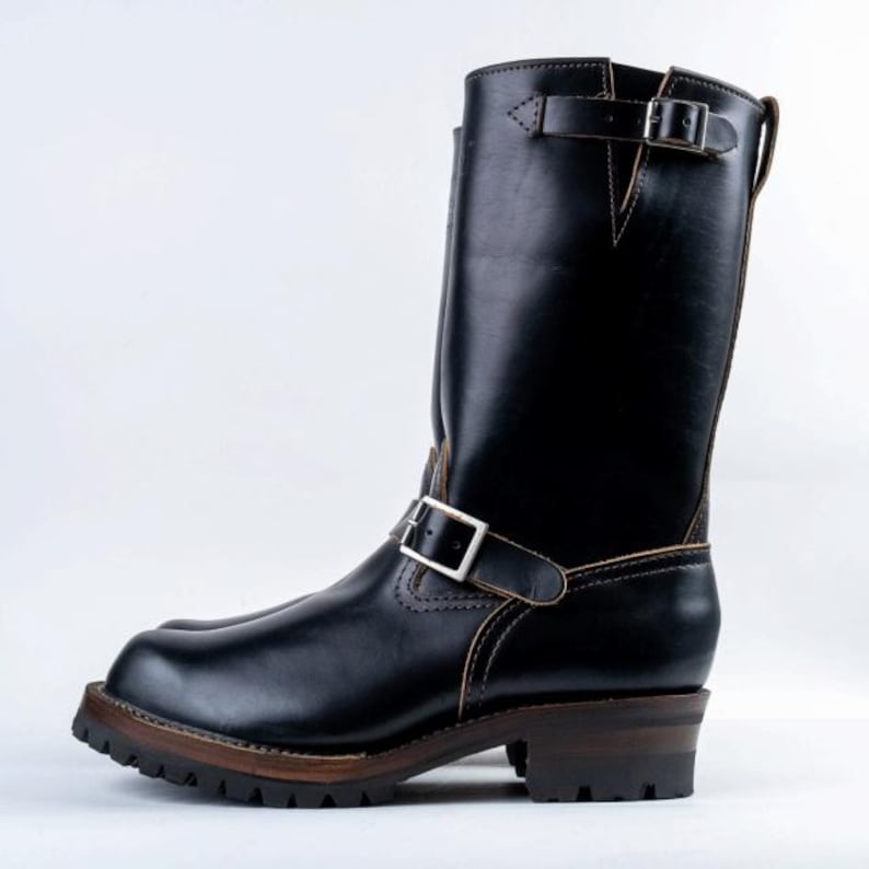 May include: Black leather motorcycle boot with a tall shaft, featuring two adjustable buckle straps. The boot has a lug sole and a stacked heel, with visible stitching details. The overall design is rugged and durable.