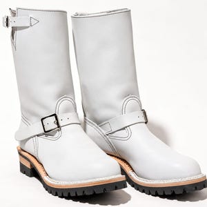 May include: A pair of white leather mid-calf boots featuring a silver-tone buckle strap across the instep and a smaller buckle at the top of the shaft. The boots have a thick black lug sole with a visible light-colored welt and white stitching.