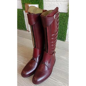 May include: A pair of knee-high burgundy leather riding boots featuring a front zip closure, a buckled strap at the top, and a secondary buckled strap across the ankle. The boots have a smooth finish with a quilted panel on the outer shaft.