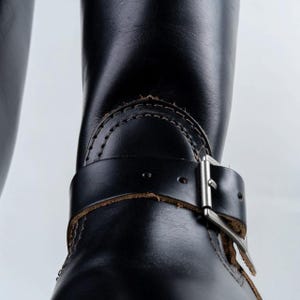 May include: Close-up of a black leather boot with a buckle and strap detail. The boot features visible stitching and a silver-colored buckle. The leather appears worn, adding character to the footwear.