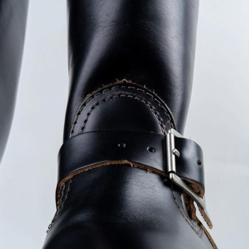 May include: Close-up of a black leather boot with a buckle and strap detail. The boot features visible stitching and a silver-colored buckle. The leather appears worn, adding character to the footwear.