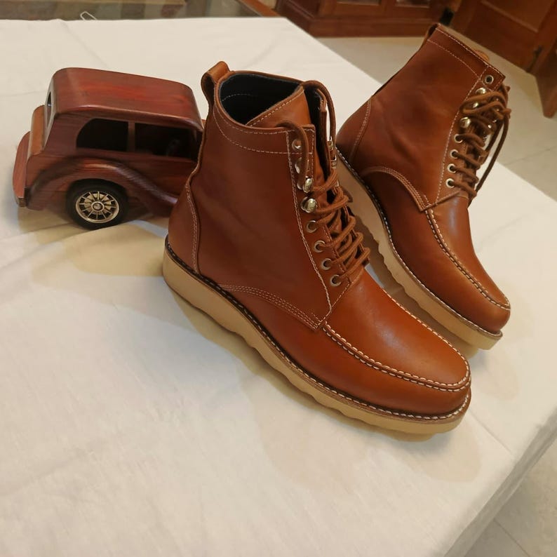 May include: A pair of brown leather boots with tan soles and white stitching. The boots have laces and metal eyelets. A wooden car model is in the background.