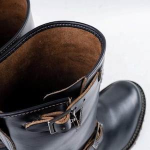 May include: Black leather motorcycle boots with brown leather interior. The boots have buckles and straps for closure and a stitched sole. The boots are designed for durability and style.