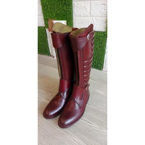 May include: A pair of burgundy leather riding boots. The boots have a tall shaft with a zip closure and buckle detail. The boots are made of smooth leather and have a classic design.