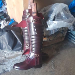 Puede incluir: Un par de botas altas de cuero burdeos con hebillas doradas y un diseño acanalado. Las botas tienen un aspecto elegante y pulido y están diseñadas para llegar a la rodilla. Las botas están de pie.