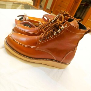 May include: A pair of brown leather work boots with white stitching and tan soles. The boots have metal eyelets and brown laces. The boots are displayed on a white surface.