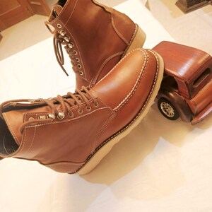 May include: A pair of brown leather boots with white stitching and laces. The boots have a classic design with a light-colored sole. A wooden toy car is next to the boots.
