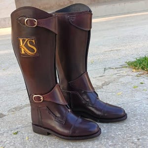 Op de afbeelding: Een paar bruine leren rijlaarzen met een gouden geborduurd "KS"-logo op de linker laars. De laarzen hebben een rits aan de achterkant en een gespsluiting aan de voorkant.