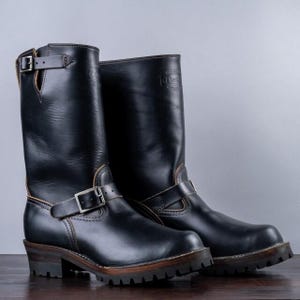 May include: A pair of black leather motorcycle boots with silver buckles and straps. The boots have a rugged sole and a mid-height shaft. The boots are displayed on a wooden surface.