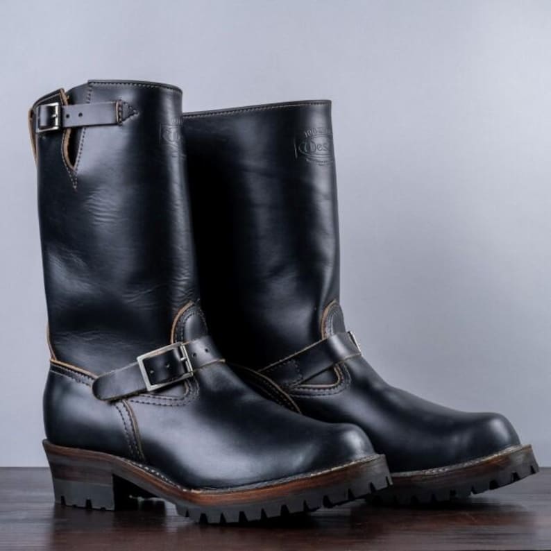 May include: A pair of black leather motorcycle boots with silver buckles and straps. The boots have a rugged sole and a mid-height shaft. The boots are displayed on a wooden surface.