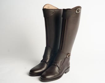 Puede incluir: Un par de botas de montar de color marrón oscuro hasta la rodilla, con cierre de cremallera frontal, punta redondeada y una correa decorativa con hebilla de metal plateado en la parte superior. Acabado liso y diseño de caña estructurada.