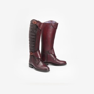 Puede incluir: Un par de botas altas de montar de cuero burdeos. Las botas tienen un diseño clásico con un acabado liso, acentuado por costuras decorativas y hebillas. Las botas miden aproximadamente 40 cm de alto.