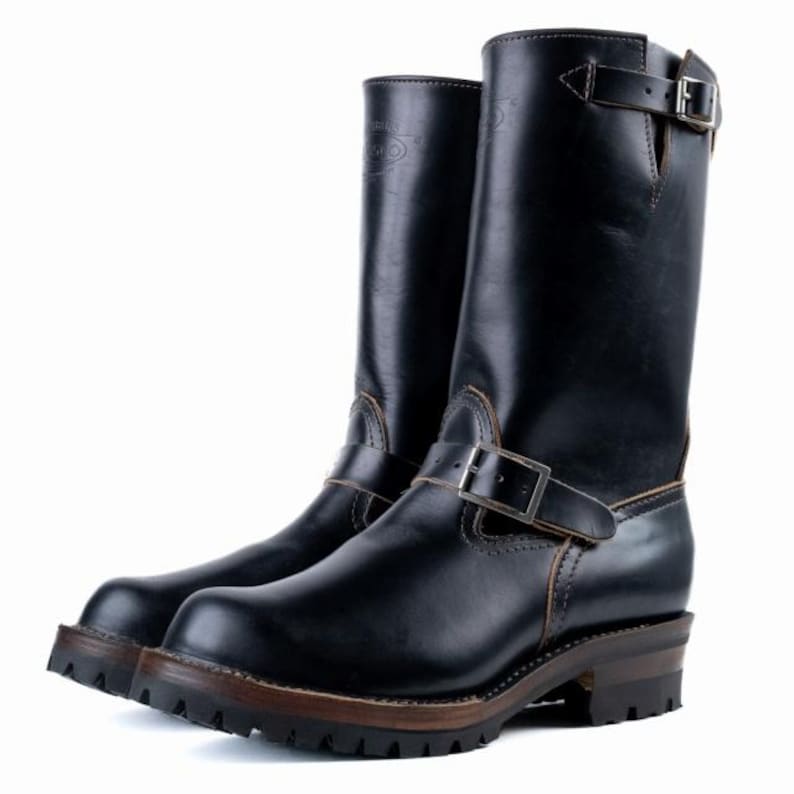 May include: Black leather motorcycle boots with a buckle closure and a lug sole. The boots have a tall shaft and a brown sole. The leather has a glossy finish. The boots are designed for durability and style.