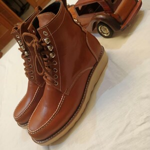 May include: A pair of brown leather ankle boots with a lace-up design and a light-colored sole. The boots feature silver eyelets and white stitching details. The boots are approximately 6 inches tall.
