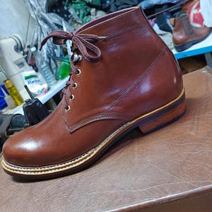 Könnte beinhalten: Ein polierter brauner Lederstiefeletten mit abgerundeter Zehenpartie, Schnürung vorne mit dunkelbraunen Schnürsenkeln, goldfarbenen Ösen und einem gestapelten Holzabsatz mit dunkler Sohle. Der Stiefel hat sichtbare Nähte an den Seitenteilen und am Rahmen.