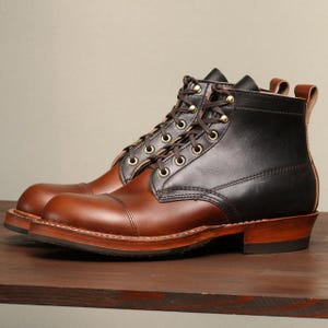 May include: A pair of two-tone leather boots. The boots are brown on the lower half and black on the upper half. They have brown laces, gold eyelets, and a wooden sole. The boots are displayed on a wooden surface.